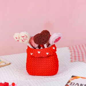 Puede incluir: Un recipiente rojo tejido con detalles de corazones blancos, que contiene varias piruletas en forma de coraz&oacute;n. Las piruletas son rosas, blancas y marrones. El recipiente est&aacute; sobre un mantel de encaje blanco, sobre un fondo rosa.