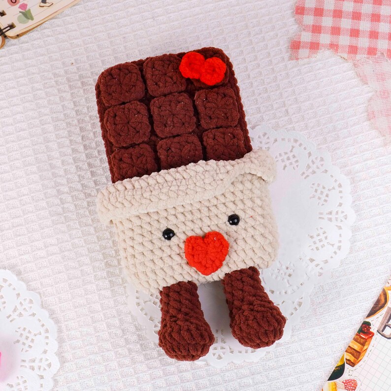 Puede incluir: Una barra de chocolate hecha a mano con ganchillo con un coraz&oacute;n rojo y un lazo. La barra de chocolate es marr&oacute;n, con un envoltorio color crema y peque&ntilde;os pies marrones. Los ojos son negros.