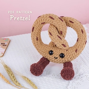 Puede incluir: Un juguete de pretzel hecho a mano, con una cara sonriente y detalles en marrón oscuro. El pretzel es de color marrón claro con manchas marrones más oscuras y tiene dos pequeñas patas marrones oscuras. El texto "PDF PATTERN Pretzel" es visible.