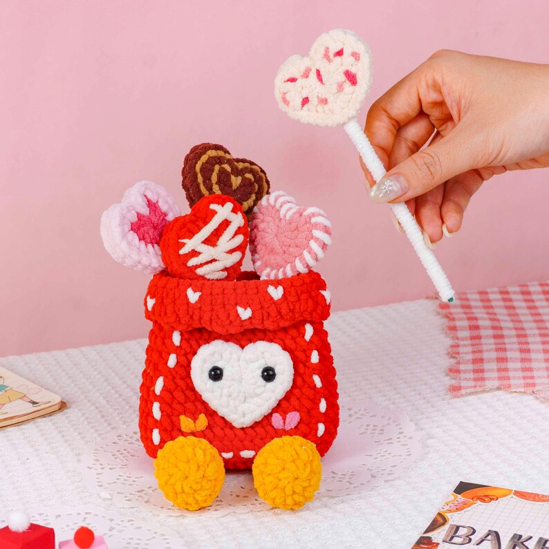 Puede incluir: Una bolsa roja tejida a ganchillo con un dise&ntilde;o de coraz&oacute;n blanco, que contiene varios caramelos en forma de coraz&oacute;n tejidos a ganchillo. Una mano sostiene una piruleta en forma de coraz&oacute;n. La bolsa tiene dos pompones amarillos en la parte inferior. El fondo es rosa.
