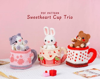 May include: Three crocheted cups with animal figures. The cups are decorated with hearts, strawberries, and paw prints. The text "Sweetheart Cup Trio" is at the top. The animals include a cat, a bunny, and a bear.