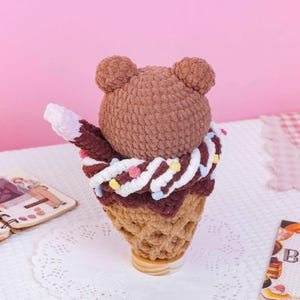 May include: A brown crocheted bear head sits atop a dark brown crocheted ice cream scoop, which is nestled in a tan crocheted waffle cone. The ice cream is decorated with a white crocheted swirl and small, colorful pom-poms in pink, yellow, and blue.