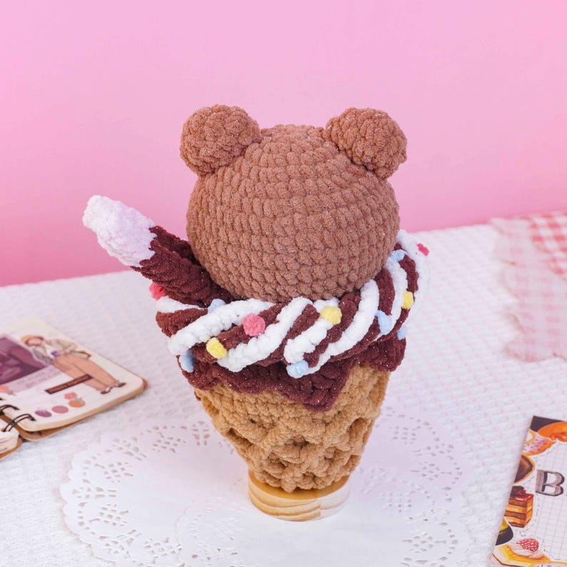 May include: A brown crocheted bear head sits atop a dark brown crocheted ice cream scoop, which is nestled in a tan crocheted waffle cone. The ice cream is decorated with a white crocheted swirl and small, colorful pom-poms in pink, yellow, and blue.