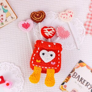 Puede incluir: Una bolsa roja de ganchillo con un coraz&oacute;n blanco y piernas amarillas, rodeada de piruletas en forma de coraz&oacute;n de ganchillo en varios colores. Las piruletas est&aacute;n sobre palitos blancos. Un peque&ntilde;o cuaderno y un letrero de panader&iacute;a est&aacute;n en el fondo.