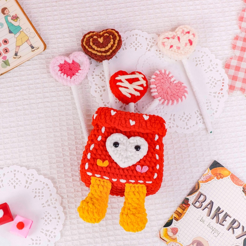 Puede incluir: Una bolsa roja de ganchillo con un coraz&oacute;n blanco y piernas amarillas, rodeada de piruletas en forma de coraz&oacute;n de ganchillo en varios colores. Las piruletas est&aacute;n sobre palitos blancos. Un peque&ntilde;o cuaderno y un letrero de panader&iacute;a est&aacute;n en el fondo.