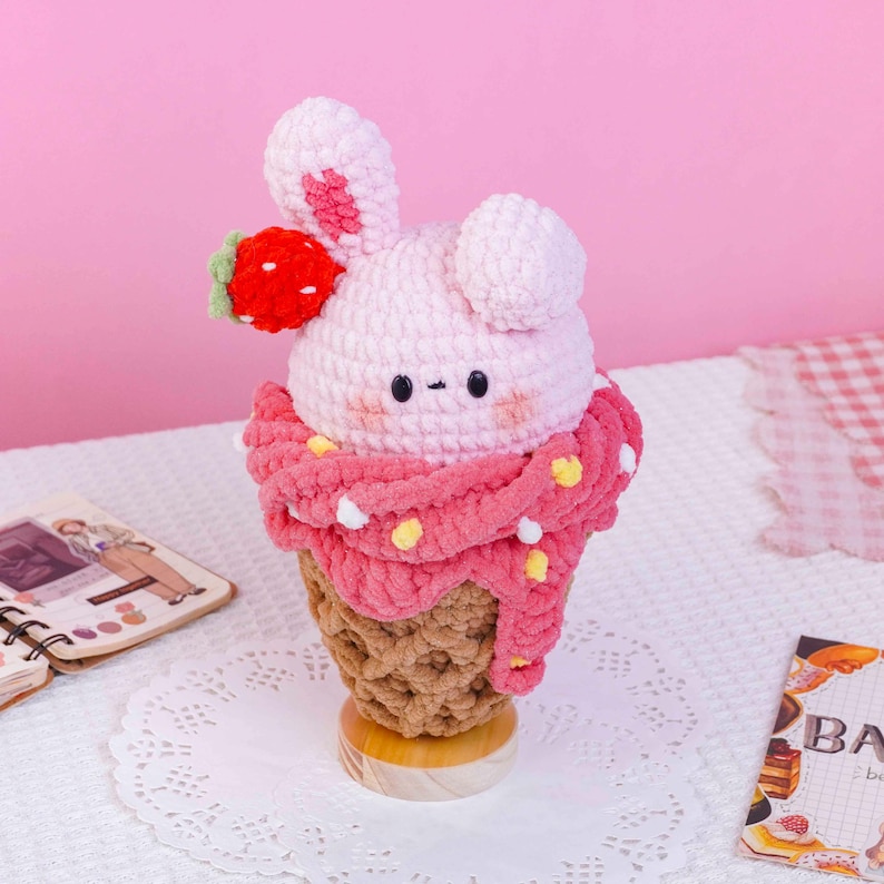 May include: A crochet plush toy shaped like an ice cream cone with a light pink bunny head featuring black eyes, a small mouth, and a red strawberry accent. The cone is brown, and the ice cream scoop is wrapped in a pink scarf with yellow dots.