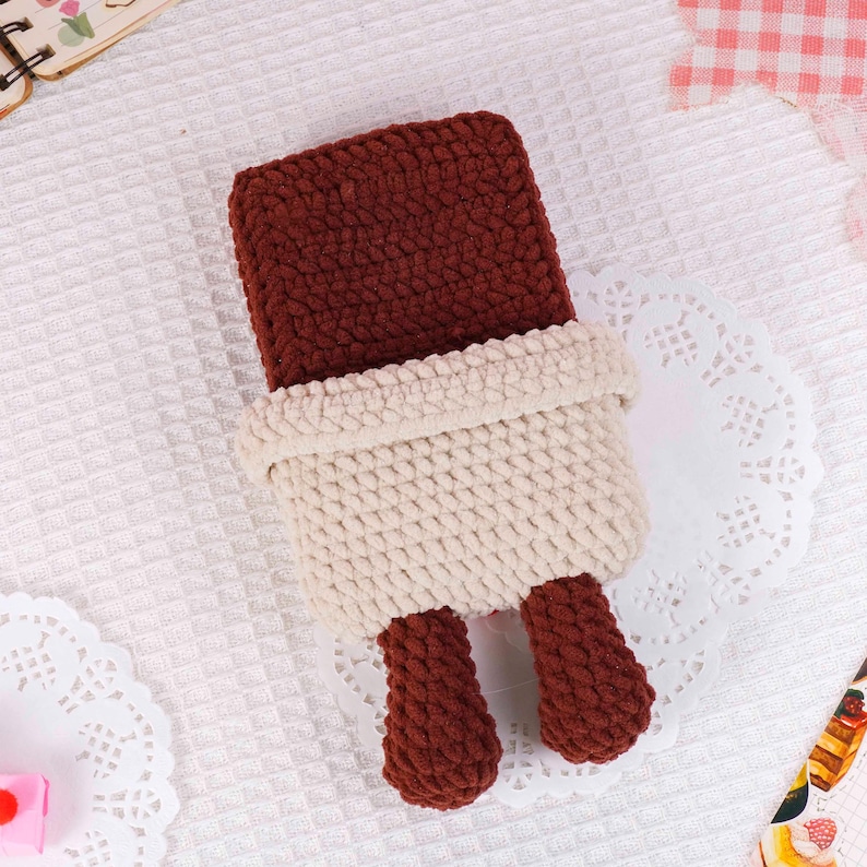 Puede incluir: Una funda de tel&eacute;fono hecha a mano con ganchillo en un dise&ntilde;o &uacute;nico. La funda presenta una parte superior rectangular de color marr&oacute;n oscuro, una secci&oacute;n media de color crema y dos peque&ntilde;os pies marrones. La funda est&aacute; colocada sobre una superficie blanca texturizada.
