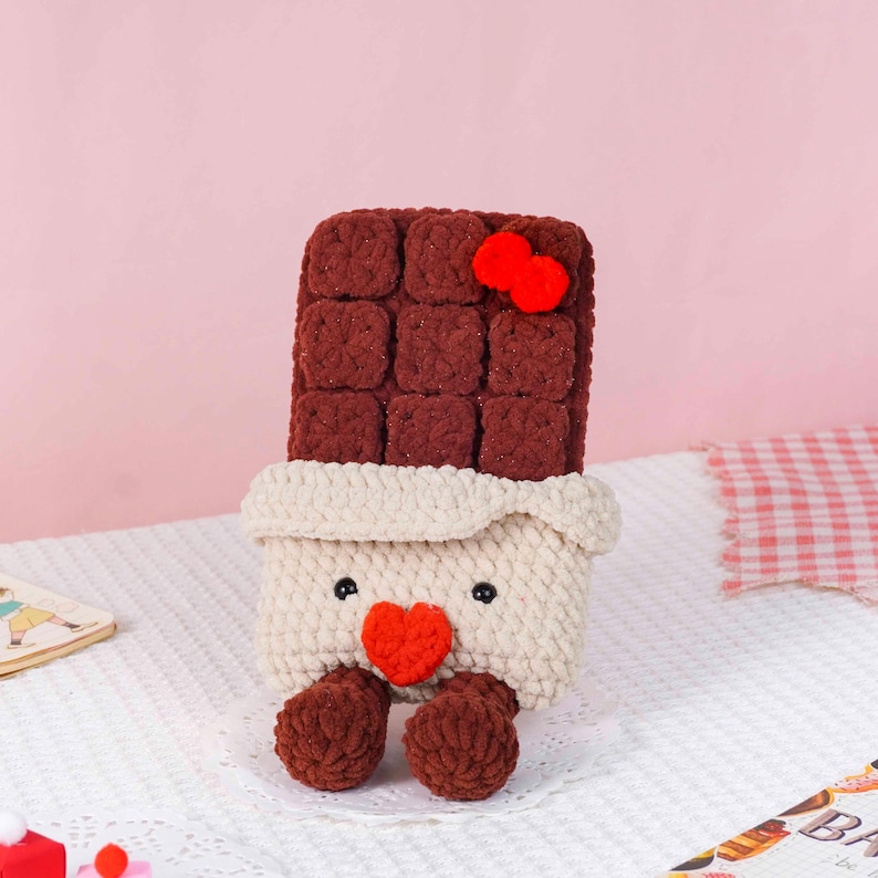 Puede incluir: Una barra de chocolate de ganchillo hecha a mano con un lazo rojo, un coraz&oacute;n y pies. La barra de chocolate es marr&oacute;n, con un envoltorio color crema y peque&ntilde;os ojos negros. El art&iacute;culo est&aacute; sobre una superficie blanca.