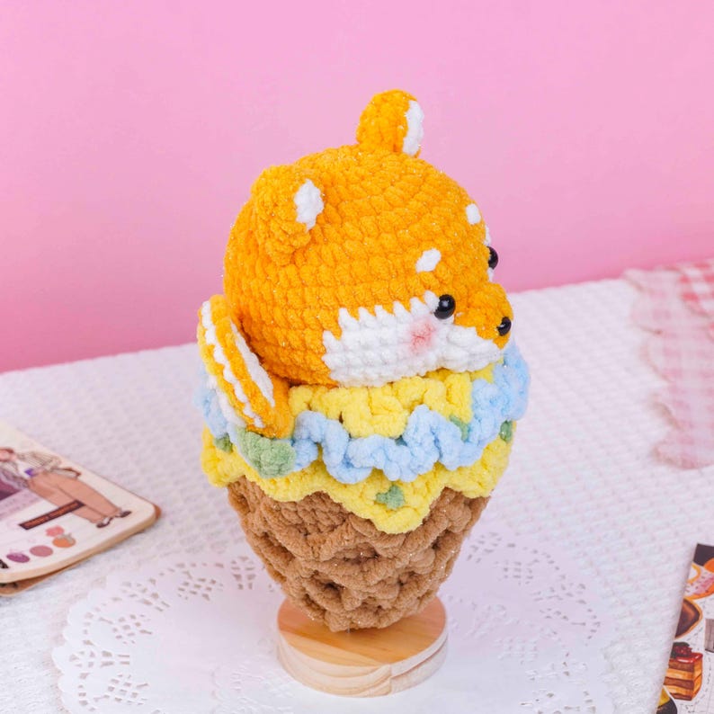 May include: A crochet plush toy shaped like a Shiba Inu dog head emerging from a brown waffle cone. The dog has orange and white yarn, black eyes, and pink cheeks. A light blue and yellow ruffled yarn trim sits between the dog and the cone base.