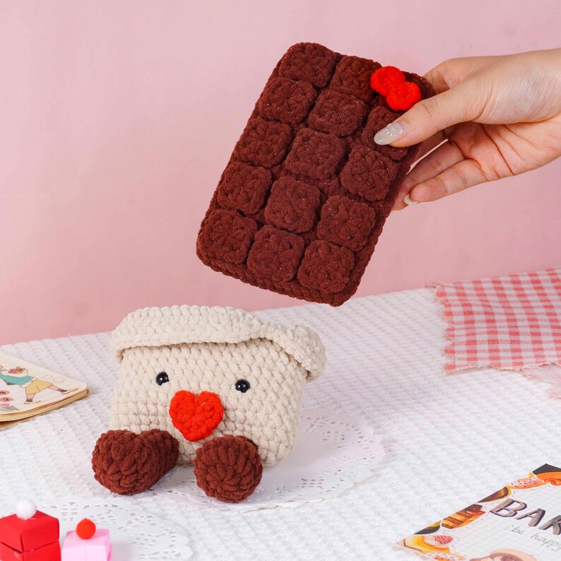 Puede incluir: Una barra de chocolate y un recipiente a juego con forma de oso, ambos hechos a mano con ganchillo. La barra de chocolate es de color marr&oacute;n oscuro con un peque&ntilde;o coraz&oacute;n rojo. El recipiente con forma de oso es de color crema con un coraz&oacute;n rojo y patas marrones.
