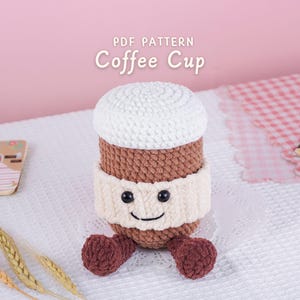 Puede incluir: Una taza de café de ganchillo con la parte superior blanca, el cuerpo marrón y la manga color crema. La taza tiene una cara sonriente con ojos negros y pequeños pies redondos de color granate. El texto "PDF PATTERN Coffee Cup" está en la parte superior.