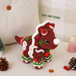 May include: A brown crocheted triceratops toy decorated with white, red, and green yarn accents resembling icing and candy. The dinosaur features a large frill, a horn, and dark eyes. The text PDF PATTERN is visible above the toy.
