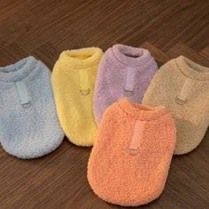 Peut inclure: Cinq pulls pour chiens aux couleurs pastel : bleu, jaune, violet, orange et beige. Chaque pull est doté d'une sangle assortie avec un anneau métallique. Les pulls sont faits d'une matière douce et duveteuse.