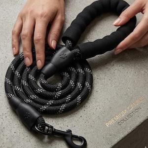 Peut inclure: Laisse pour chien à motifs noirs et blancs avec une poignée rembourrée. La laisse est enroulée et comporte un mousqueton noir et des détails de pattes. Le texte "PREMIUM CANINE COLLECTION" est visible.