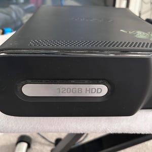 Puede incluir: Consola Xbox 360 negra con una etiqueta de 120 GB HDD. La consola tiene una ventilación en la parte superior y una pegatina verde en el lateral. La consola está sobre una superficie blanca.