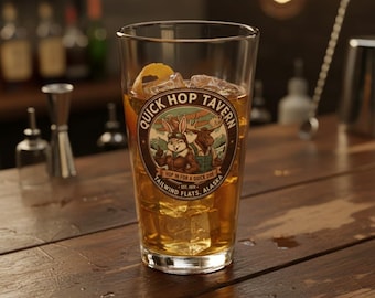 May include: A tall glass filled with ice and a golden-colored beverage, garnished with an orange peel. The glass features a circular emblem with a rabbit and moose, and the text "Quick Hop Tavern, Tailwind Flats, Alaska."