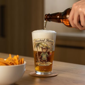 Peut inclure: Un verre transparent rempli de bière dorée et de mousse blanche, orné d'un singe portant un chapeau de cowboy et une chemise à fleurs sur une plage. Le texte Mischief Maker et Pleasure Taker est imprimé sur le verre. La bière est versée d'une bouteille brune.