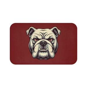 May include: A rectangular bath mat featuring a detailed illustration of a bulldog's head. The bulldog has a stern expression with red eyes and a cream-colored face. The background is a solid maroon color.