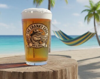 May include: A pint glass filled with beer and a foamy head, featuring "The Cranky Croc Bar & Grill" logo. The logo depicts an alligator wearing a hat and smoking a pipe. The glass sits on a wooden surface with a beach and palm trees in the background.
