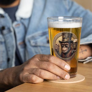 May include: A pint glass filled with beer, featuring a circular graphic of a chimpanzee wearing a top hat and holding a beer. The graphic includes the text "BATTERED HAT BREWING". The glass is resting on a cork coaster.