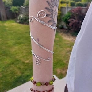 May include: A silver arm cuff with a decorative swirl design, worn on the upper arm. Two beaded bracelets in green and brown are on the wrist. A sun tattoo is visible. The background shows a green lawn and trees.