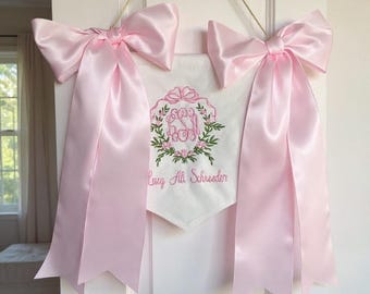 May include: A white banner with pink satin bows and ribbons. The banner features an embroidered floral wreath with initials and the name "Lucy Ali Schrester". The banner hangs from a gold cord.