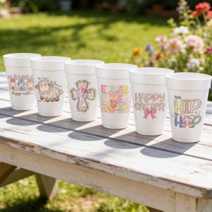 May include: Six white foam cups with Easter-themed designs. The cups feature images of bunnies, a lamb, a cross, and the phrases "Happy Easter" and "Too Hip to Hop." The cups are arranged on a weathered wooden table outdoors.