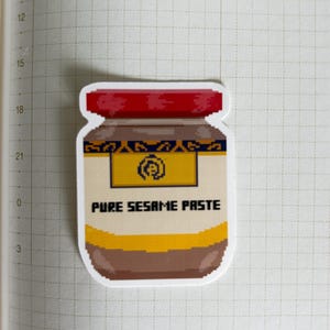 May include: A sticker featuring a pixelated jar of pure sesame paste. The jar has a red lid, a yellow label with a symbol, and the words "PURE SESAME PASTE" in black. The jar's body is brown and beige.