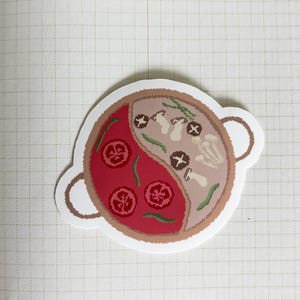 May include: A white sticker with a red and brown illustration of a hot pot with vegetables and mushrooms. The hot pot is divided in half, with one side showing red broth and the other side showing a lighter broth.
