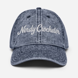 May include: A denim blue baseball cap with a curved brim. The cap features the embroidered text "Nerdy Crocheter" in white script across the front. The hat has a vintage, worn look.