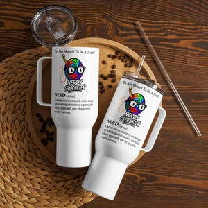 May include: Two white travel mugs with the text "Nerdy Crocheter" and a colorful yarn ball design with glasses. One mug has a lid and the other has a straw. The mugs are on a wooden tray with coffee beans.