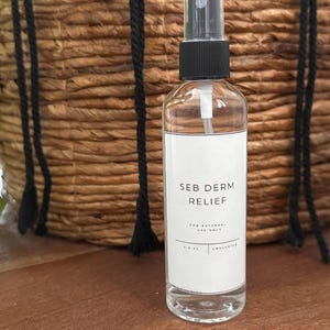 May include: A clear glass bottle with a black spray top and a white label that reads "SEB DERM RELIEF" and "FOR EXTERNAL USE ONLY" in black text. The bottle is sitting on a wooden surface.