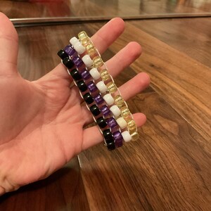May include: Four beaded bracelets in black, purple, white, and gold. Each bracelet is made of small, round beads strung on a white cord. The bracelets are held in a hand, showcasing the colours and design.