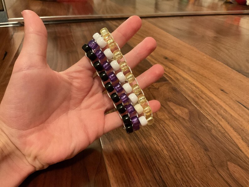 May include: Four beaded bracelets in black, purple, white, and gold. Each bracelet is made of small, round beads strung on a white cord. The bracelets are held in a hand, showcasing the colours and design.