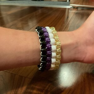 May include: Four beaded bracelets in black, purple, white, and gold are stacked on a wrist. The bracelets are made of small, round beads strung together. The wrist is resting on a wooden surface.