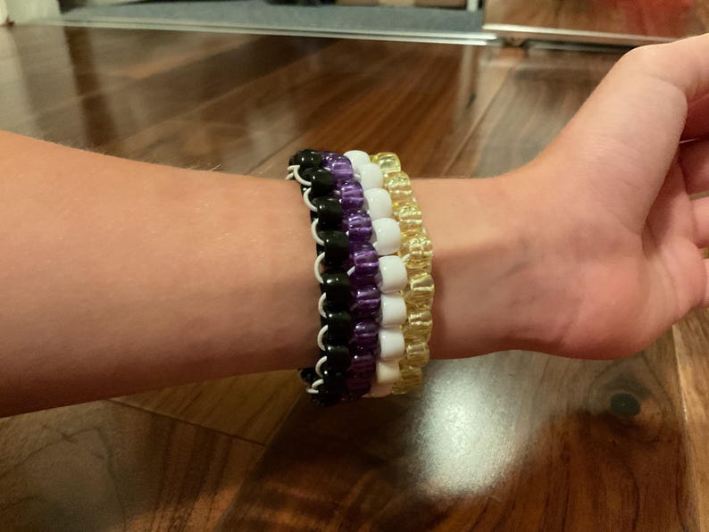 May include: Four beaded bracelets in black, purple, white, and gold are stacked on a wrist. The bracelets are made of small, round beads strung together. The wrist is resting on a wooden surface.