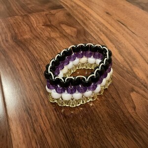 May include: A beaded bracelet with a layered design. The bracelet features black, purple, white, and gold beads. The beads are strung together to form a thick, circular band. The bracelet is resting on a wooden surface.