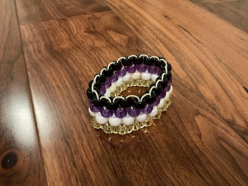 May include: A multi-layered beaded bracelet in black, purple, white, and gold. The beads are arranged in a circular pattern. The bracelet is displayed on a wooden surface. The bracelet's design is eye-catching.