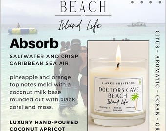May include: A lit candle in a clear glass jar with a label that reads "Doctors Cave Beach Island Life." The candle is made with luxury hand-poured coconut apricot wax and has a fresh air, lively scent. The image includes text about the scent profile.