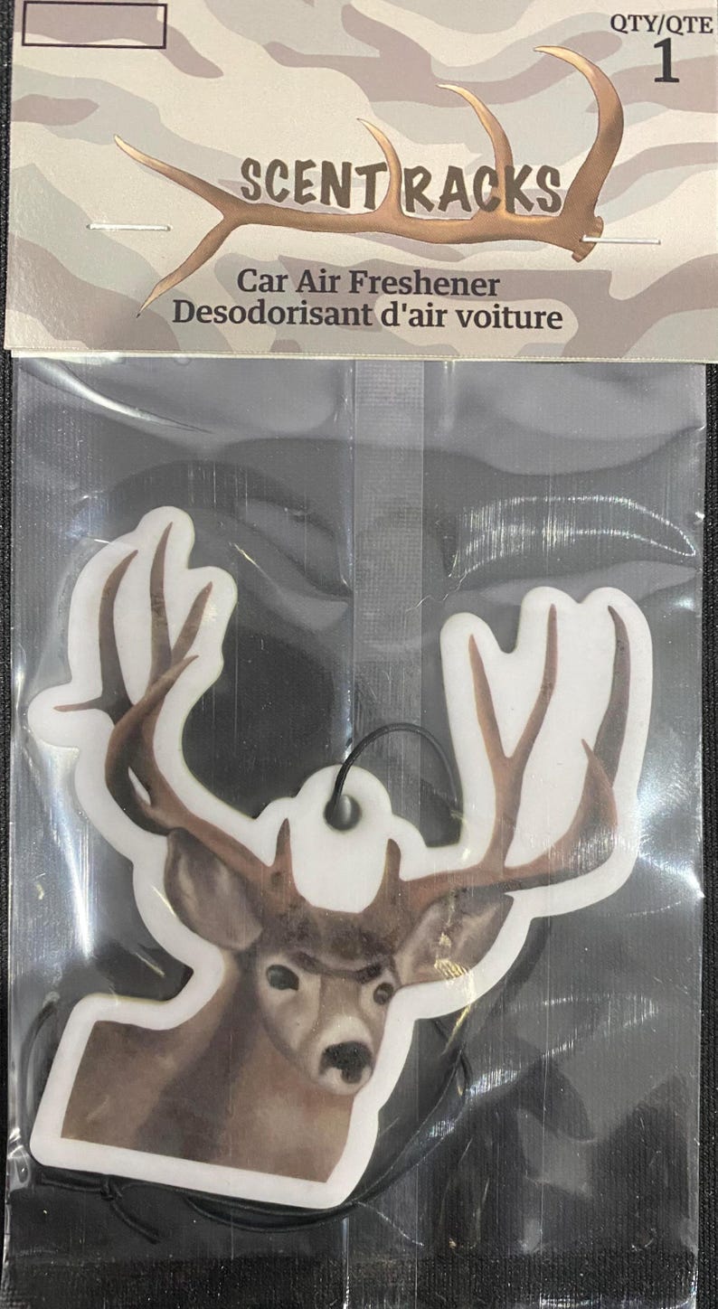 May include: A white car air freshener with a brown deer head and antlers. The text "SCENT RACKS" is printed on the packaging. The text "Car Air Freshener" and "Desodorisant d'air voiture" is printed on the packaging. The packaging is camo-patterned.