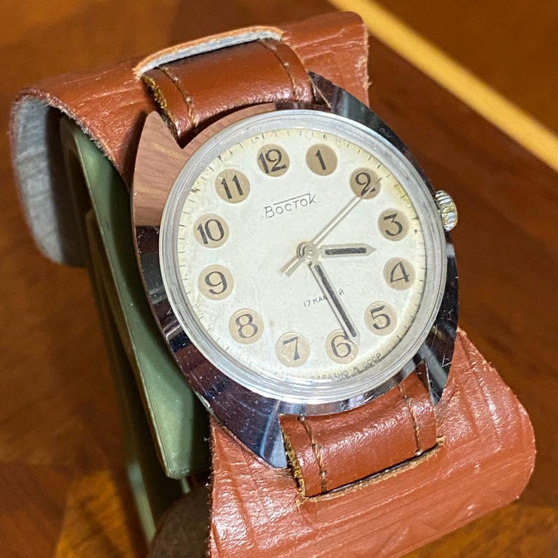Może przedstawiać: Zegarek Vostok w stylu vintage z kremową tarczą, czarnymi cyframi i srebrnymi wskaz&oacute;wkami. Zegarek ma kopertę ze stali nierdzewnej i brązowy sk&oacute;rzany pasek. Zegarek jest prezentowany na brązowym sk&oacute;rzanym uchwycie.