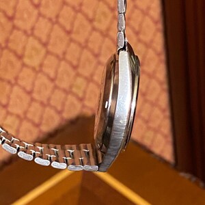 May include: A silver-toned wristwatch with a metallic band. The watch face is a light brown color. The watch is hanging in front of a patterned orange background.