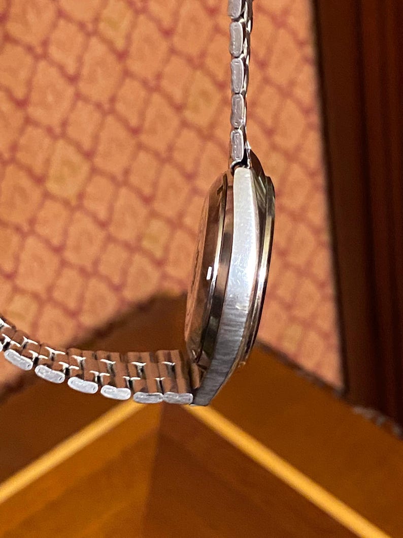 May include: A silver-toned wristwatch with a metallic band. The watch face is a light brown color. The watch is hanging in front of a patterned orange background.