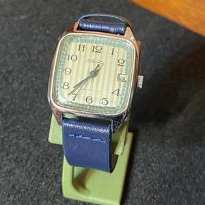 May include: A vintage wristwatch with a rectangular gold-toned case and a striped, light green and yellow face. The watch has black numerals and hands, a date window, and a blue leather strap. The watch is displayed on a green stand.