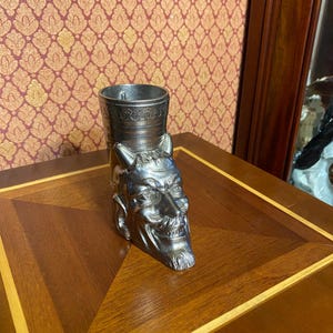Puede incluir: Un vaso de chupito de color plateado con un diseño de cara de diablo. El vaso tiene forma cilíndrica con una representación detallada de la cara de un diablo en la parte delantera, con cuernos y perilla. El vaso de chupito es de metal.