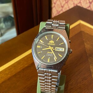 May include: A silver-toned Orient watch with a dark brown face, gold hands, and markers. The watch displays the day and date. The watch is on a green stand, with a silver band. The watch face has the text "Orient" and "Crystal 21 Jewels".