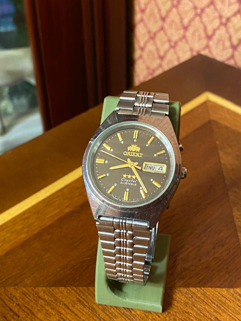 May include: A silver-toned Orient watch with a dark brown face, gold hands, and markers. The watch displays the day and date. The watch is on a green stand, with a silver band. The watch face has the text "Orient" and "Crystal 21 Jewels".