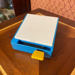May include: A blue and white box with a white lid featuring a detailed embossed design of a building. A yellow square button is on the side of the box. The box is on a wooden surface.