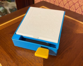 May include: A blue and white box with a white lid featuring a detailed embossed design of a building. A yellow square button is on the side of the box. The box is on a wooden surface.