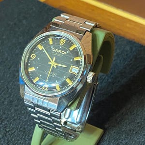 Puede incluir: Un reloj de pulsera automático Cardi vintage con una correa de acero inoxidable y una esfera negra. El reloj tiene manecillas e indicadores de hora dorados, y la esfera muestra el logotipo de Cardi y la palabra "Crystal". El reloj se exhibe en un soporte verde.